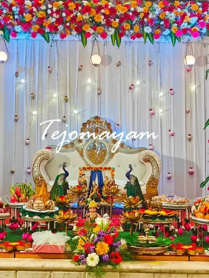 A full stage view of a baby shower decoration, featuring a dense floral arch and a long table filled with traditional offerings and dolls.