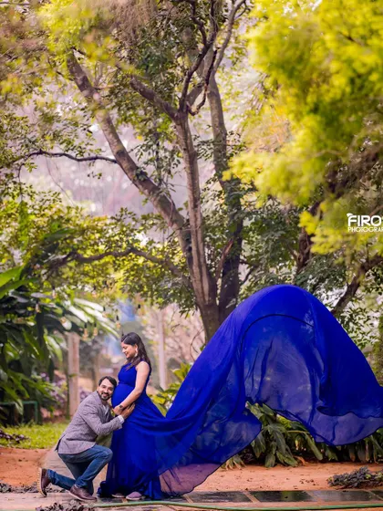 A beautiful portrait of an expecting couple in a forest setting. The partner kneels to embrace the baby bump, while the vibrant blue gown flows out, creating a striking contrast with the natural green surroundings.