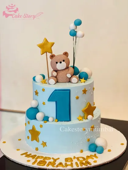 A lovely two-tier cake for a first birthday. This blue cake is decorated with stars, balloons, and a cute fondant teddy bear sitting on top.