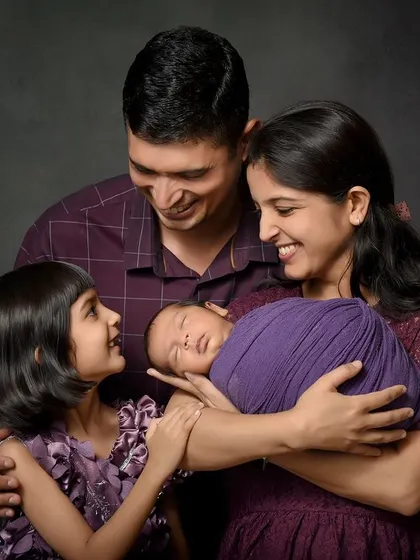 A beautiful family of four admiring their newest member. The older sister gently touches the newborn, wrapped in a vibrant purple swaddle, creating a colorful and heartwarming portrait against a dark canvas.