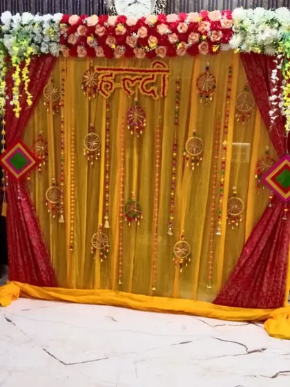 This Haldi backdrop uses a combination of sheer yellow and rich red drapes for a royal look. The decor is completed with floral arrangements at the top, a "Haldi" sign, and decorative hangings for a festive feel.