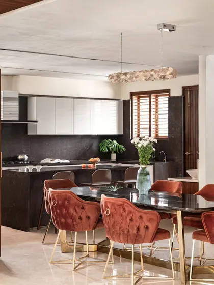 This open kitchen and dining area is designed for entertaining. The dark stone countertop extends to create a breakfast bar with seating. The rust-colored velvet chairs add a pop of color and a touch of luxury to the dining space.