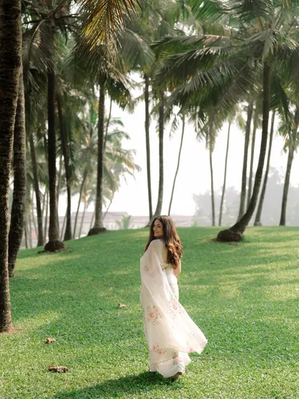 The bride looks back over her shoulder with a smile, her saree flowing as she walks through a grove of palm trees in Goa.