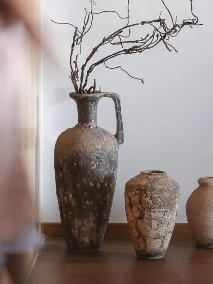 A trio of ancient-looking pottery, including a large amphora with a handle, is styled with dried branches. These pieces carry the weight of time in their texture and gentle imperfections.