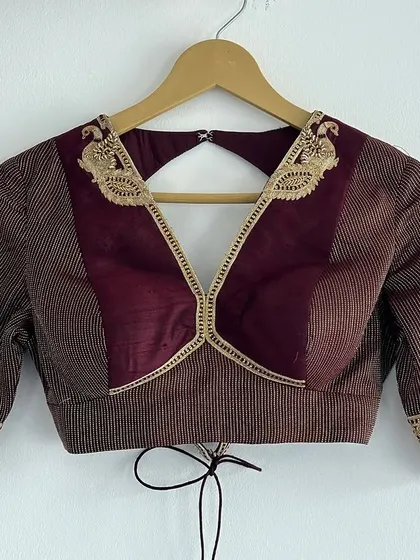 A beautifully tailored blouse in maroon and brown, featuring unique paneling and delicate peacock embroidery on the shoulders and sleeve cuffs. The V-neck design and tie-back detail add a touch of modern flair.