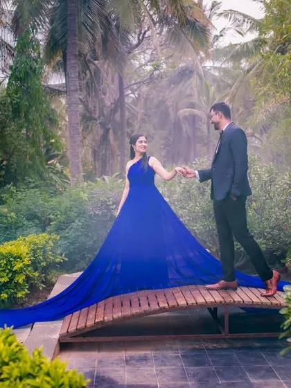 Creating a misty, dream-like atmosphere in a garden setting. The couple walks across a small wooden bridge, the long blue gown trailing behind them, for a truly magical pre-wedding photo.