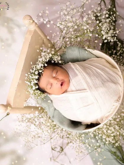 A newborn baby, wearing a floral crown, is swaddled in white and rests in a tiny bed surrounded by baby's breath. This overhead shot creates a beautiful, nest-like composition.
