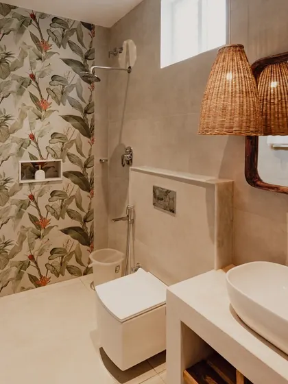 The bathroom design incorporates a tropical-themed wallpaper as a feature wall in the shower area. This brings a splash of color and nature indoors, complemented by the simple vanity and woven light pendants.