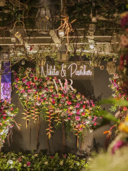 A close-up of the neon sign and floral backdrop. The cascading tropical flowers and rustic branches create a rich, textured, and modern photo opportunity.