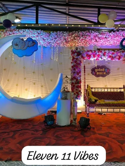 A dreamy baby shower setup featuring a large, illuminated crescent moon prop. The stage also includes a traditional swing (jhoola) decorated with a floral peacock design, blending modern and classic elements.
