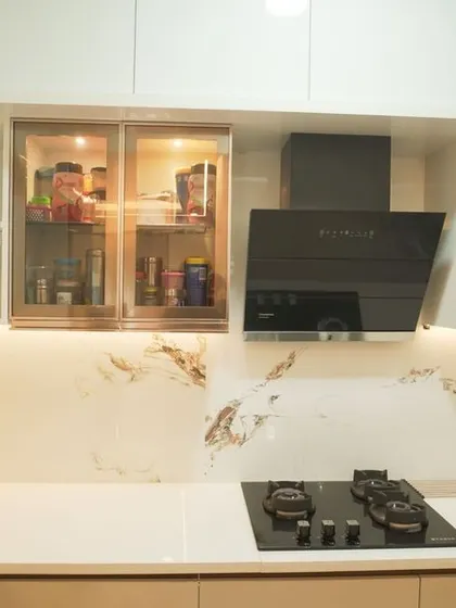 An L-shaped kitchen featuring glossy white cabinets and a quartz countertop with delicate veining. The design includes glass-fronted upper cabinets with gold-tone frames, adding a touch of elegance.