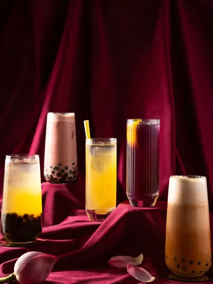 A lineup of various iced teas and bubble teas from Nara Thai. The moody lighting and red velvet background give these casual drinks a sophisticated and dramatic presentation.