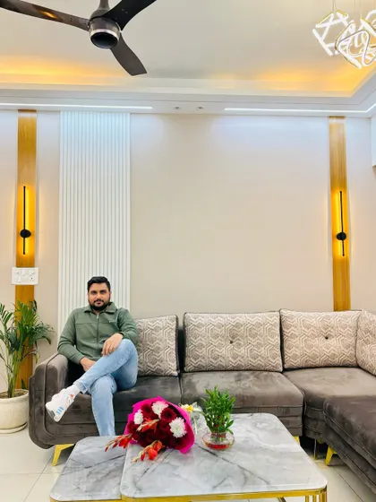 Posing in a finished living room at a Nirala Estate project. The design features a comfortable grey sofa, a marble-top table with gold accents, and elegant wall panels with integrated vertical lights.