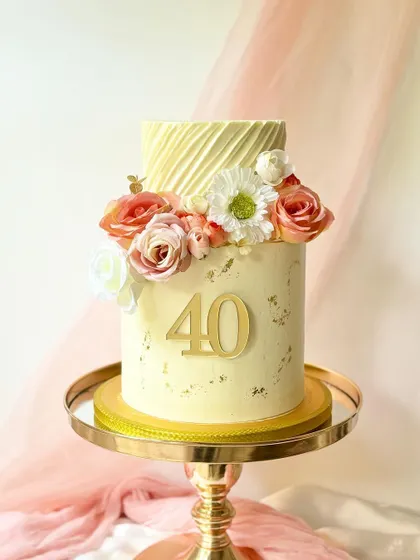 A lovely two-tier 40th birthday cake with a textured buttercream finish and gold leaf details. The cake is topped with a cascade of peach and white roses.
