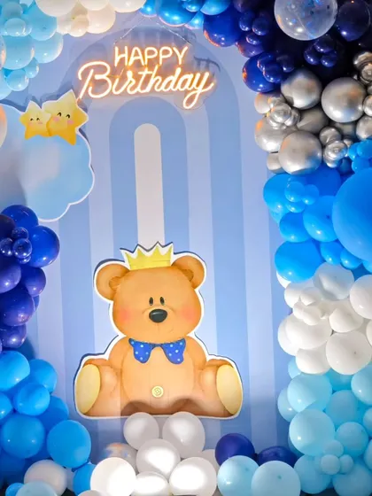 A close-up of a royal teddy bear theme, featuring a cute bear with a crown against a blue backdrop. The 'Happy Birthday' neon sign adds a modern, celebratory touch.