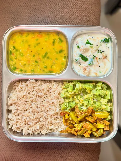 A colorful vegetarian thali with brown rice, mixed vegetable dal, sautéed snake gourd and ivy gourd, and a refreshing raita.