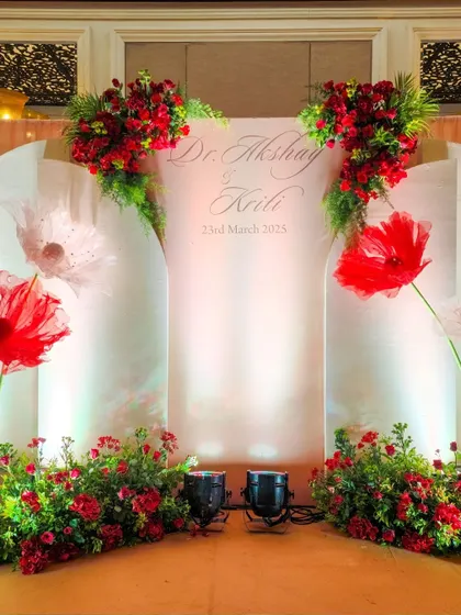 A personalized photo backdrop with the couple's name and wedding date, decorated with large, artistic paper flowers and fresh red floral arrangements.