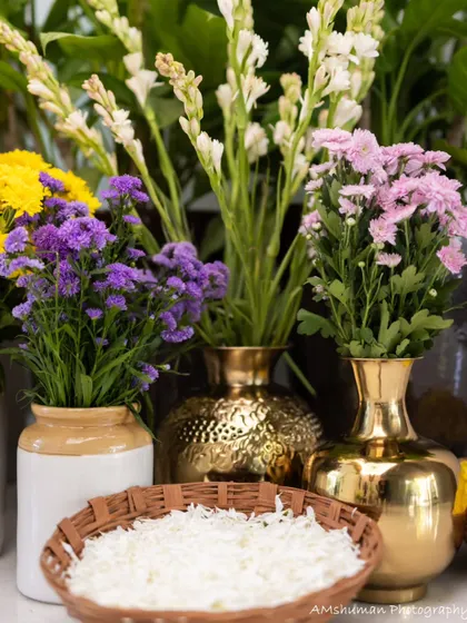A collection of fresh flowers, including tuberoses and chrysanthemums, in brass and ceramic vases, ready to be arranged for the ceremony. A bowl of jasmine petals adds a fragrant touch.