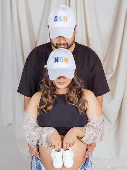 A fun and modern way to announce your new roles. This couple wears matching "Mom" and "Dad" caps, with the mom-to-be holding a tiny pair of baby shoes over her bump for a playful and casual maternity photo.