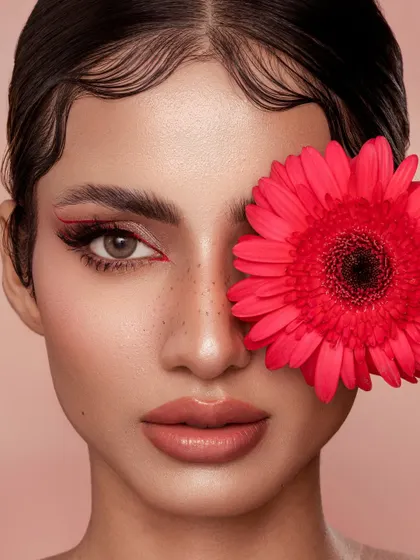 A direct, face-forward composition that emphasizes the symmetry of the look. The flower covers one eye, drawing all attention to the other, with its precise red wing and the model's freckled skin.