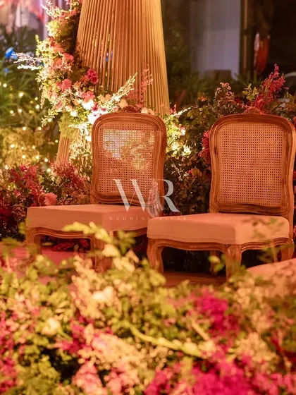 The seating for the couple at the pink floral tree mandap. The classic cane chairs are set against a backdrop of fairy lights and dense foliage, creating a romantic and intimate focal point.