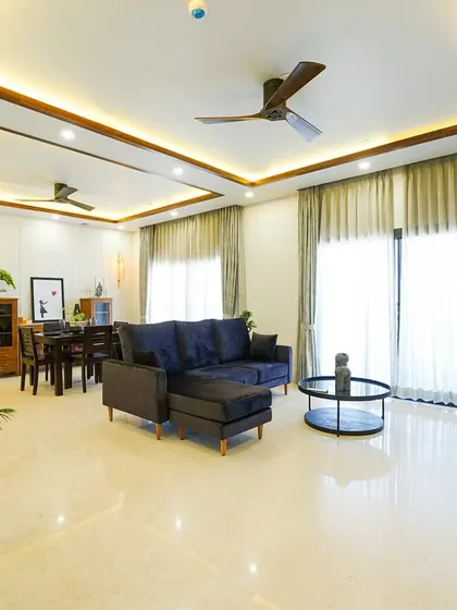 A cozy living and dining space designed for festive gatherings, featuring a deep blue sectional sofa, warm lighting from a false ceiling, and ample natural light from large windows.