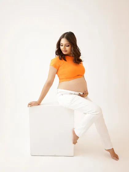 A modern and casual solo portrait. The mom-to-be shows off her bump in a bright orange crop top and white pants, creating a fresh and vibrant look.