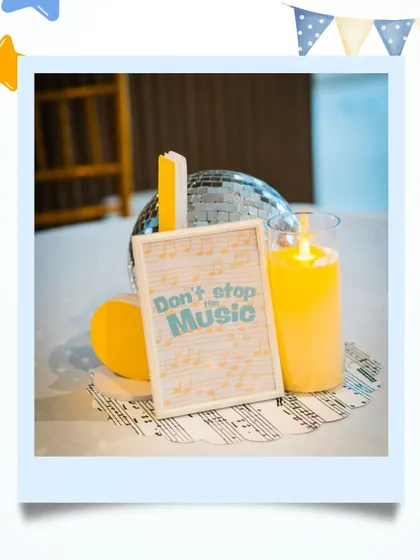 A close-up of a table centerpiece at the music-themed party, featuring a disco ball, a framed "Don't Stop the Music" sign, and a candle, all set on a musical sheet placemat.