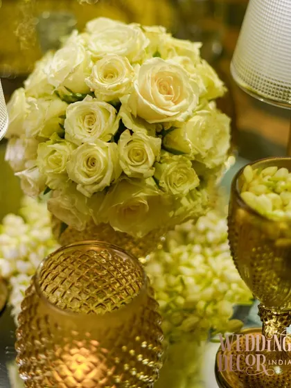 A close-up of the white rose bouquet, surrounded by the warm glow of textured gold candle holders. This highlights the romantic and classic elements of the tablescape.