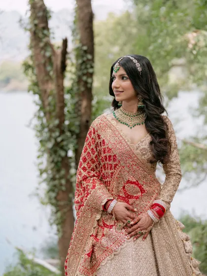 A stunning bridal portrait by the lake, her traditional red and gold attire contrasting beautifully with the natural green and blue background.