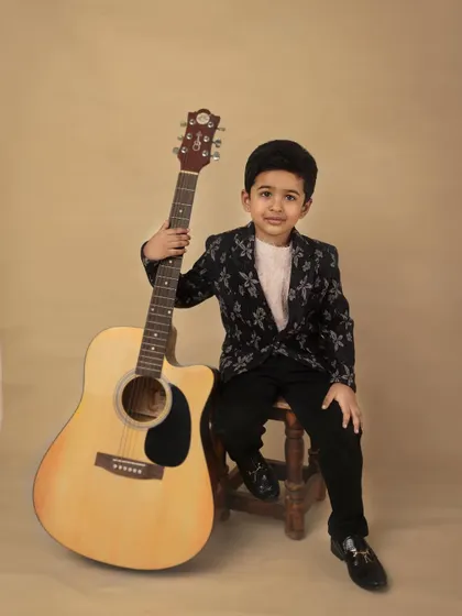 A full-length portrait of the young boy with his guitar. The warm, neutral backdrop keeps the focus entirely on him and his confident pose.