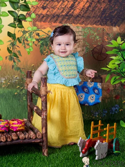 A cute baby girl in a yellow skirt and blue top, standing in a lovely village-themed setup. The vibrant colors of her outfit and the backdrop make for a cheerful and bright photo.