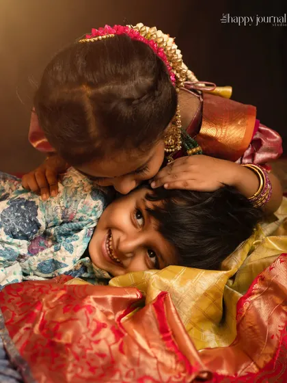 A tender moment between two siblings in traditional wear. The older sister gives a loving kiss to her smiling brother, a beautiful capture of their bond.