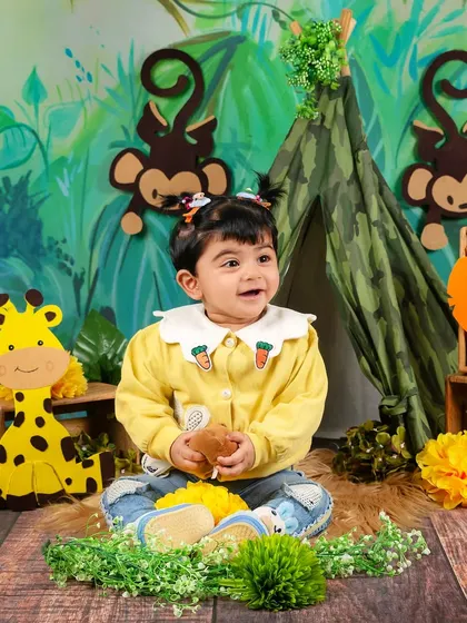 A happy baby girl in a yellow sweater enjoys her jungle-themed photoshoot, surrounded by playful animal cutouts.