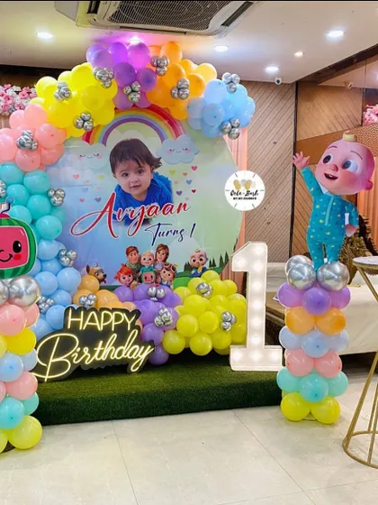 A colorful Cocomelon first birthday stage setup. It features a custom backdrop with the child's photo, a pastel balloon arch, a light-up number '1', and a Cocomelon character cutout.