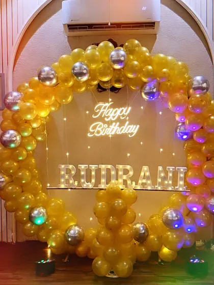 A bright and cheerful birthday setup with a yellow balloon ring, silver disco balls, a neon sign, and light-up letters for the name "Rudrani".