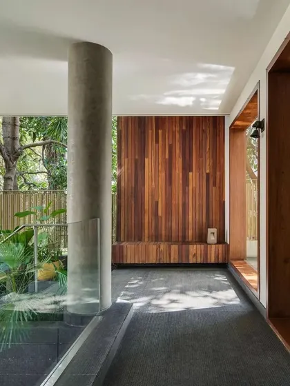 A corridor in Kanasu where raw concrete pillars and teak wood paneling meet, with sunlight filtering in to create a serene passage that feels connected to the garden.