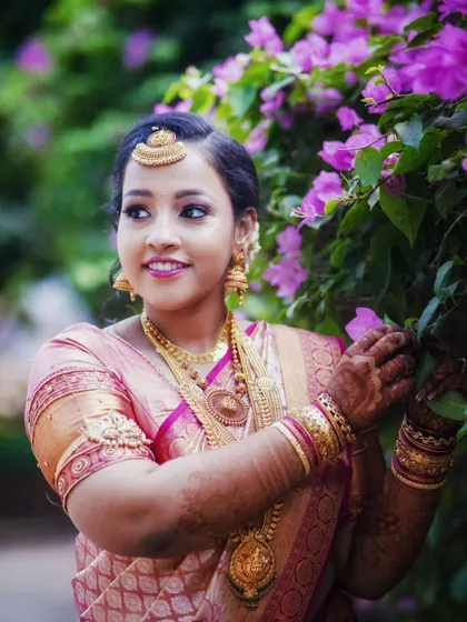 The bride poses gracefully amidst bougainvillea flowers, her traditional pink and gold saree creating a beautiful contrast.