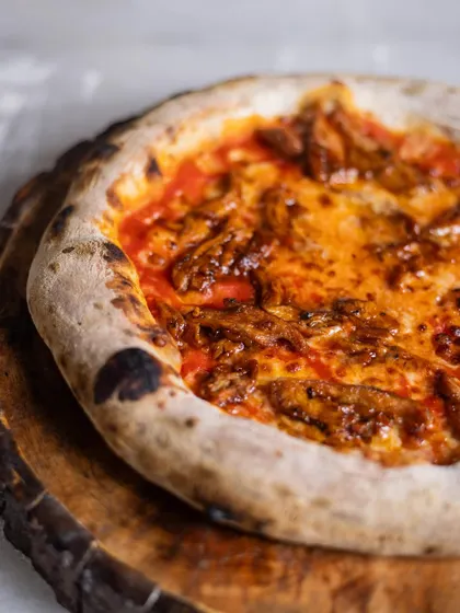 A rustic, meaty pizza sits on a wooden board. The simple presentation lets the quality of the ingredients and the perfectly charred crust speak for itself.
