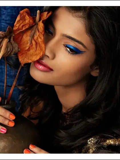 A rich, warm beauty portrait with a bold blue winged eyeliner. The use of a dried leaf as a prop adds an organic, textural element that contrasts beautifully with the sharp makeup.