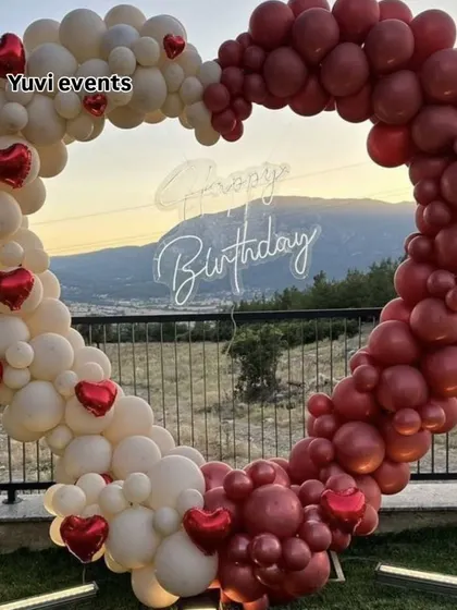 This heart-shaped balloon arch is perfect for outdoor events. The clean design with white and maroon balloons and a 'Happy Birthday' neon sign stands out beautifully against a natural background, creating a great spot for photos.