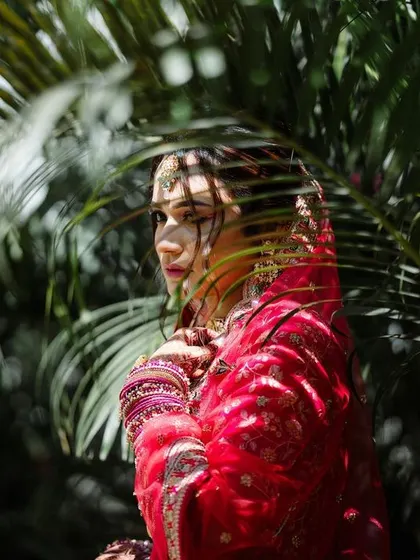 A bride in her red wedding attire, framed by palm leaves. The play of light and shadow adds a dynamic and artistic quality to this portrait.
