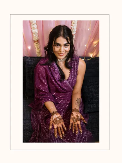 An intimate at-home Mehendi ceremony. The bride, in a stunning purple outfit, shows off her freshly applied henna, her smile lit by the warm glow of fairy lights. Home ceremonies have a special, cozy charm.