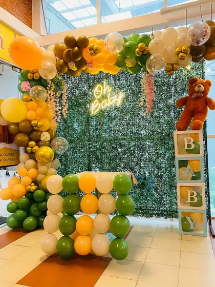 A jungle-themed "Oh Baby" baby shower. The setup features a green foliage backdrop, a balloon arch in earthy tones, and a cute teddy bear prop with "BABY" blocks.
