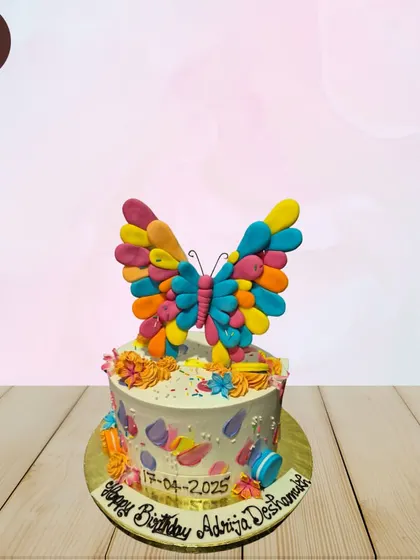 A simple and colorful butterfly cake. The main feature is a large, multi-colored butterfly topper made of individual, vibrant petals.