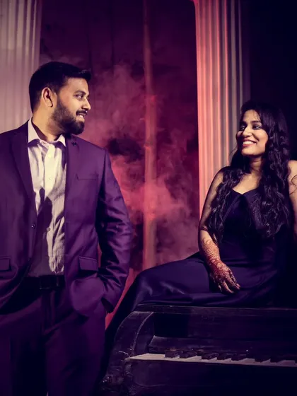 A dramatic and moody pre-wedding portrait. The groom stands beside a piano where the bride is seated, surrounded by theatrical smoke and purple lighting for a cinematic effect.