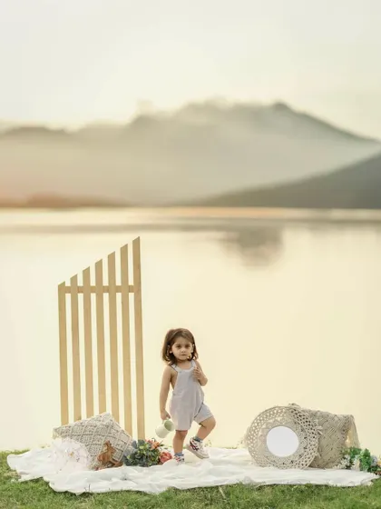 A toddler stands on a picnic blanket by a lake at sunset. The warm light, calm water, and distant mountains create a stunning backdrop for this beautiful and peaceful milestone portrait.