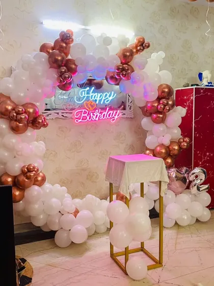 A classy rose gold and white balloon ring backdrop. This setup, complete with a neon sign and a pedestal for the cake, is perfect for an elegant birthday celebration at home.