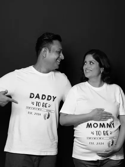 A joyful black and white photo of a couple wearing "Daddy to be" and "Mommy to be" t-shirts. Their interaction and smiles make this a heartwarming and fun shot.