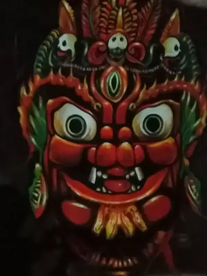 A painting of a red-faced deity mask with wide eyes and intricate headgear. The dark background makes the vibrant colors and expressive features of the mask stand out.